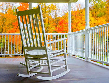 White rocking chair on a covered porch with colorful fall foliage and autumn trees in the background.
