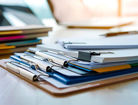 Stack of binders, notebooks, and paperwork on a desk in a bright office workspace.